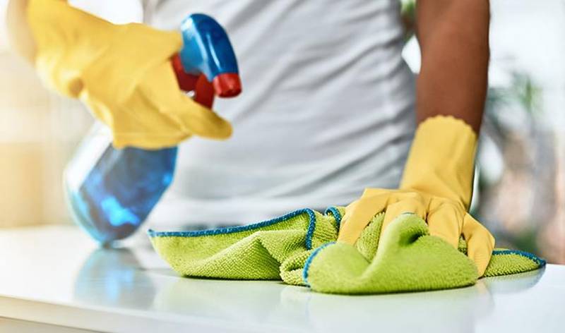 Man in yellow gloves with spray bottle in one hand and green cloth in second hand spraying on table