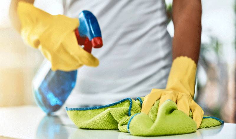 Woman in yellow gloves holding spray bottle and green cloth in hand spraying on a surface