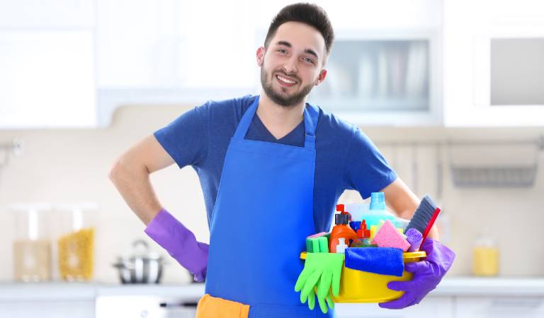 Man in blue dress holding yellow basket filled with tools, brush and chemicals.