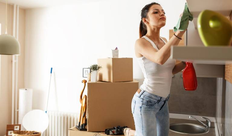 Woman in white top and jeans scrubbing cabinet with green cloth holding red bottle in her hand