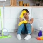 tired young black woman in rubber gloves sits on floor with mop cleaning supplies