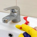 Hand in yellow rubber gloves holding a spray liquid cleaning agent for washing dirty faucet with limescale in bathroom, house cleaning concept