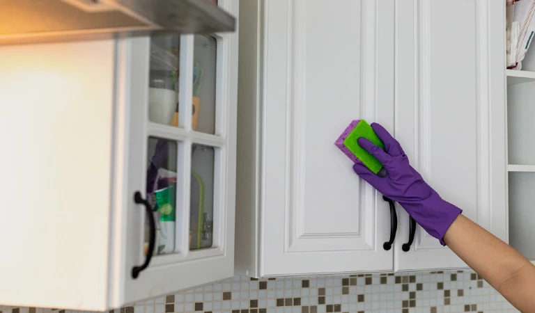 Hand in a purple rubber glove cleaning white kitchen cabinet