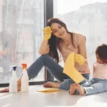 Woman with her daughter sits on window sill with bottles of cleaning spray and with gloves
