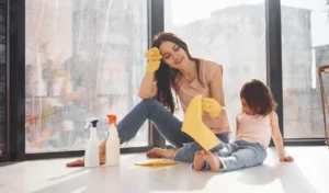 Woman with her daughter sits on window sill with bottles of cleaning spray and with gloves
