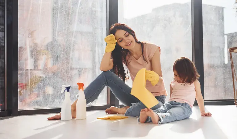 Woman with her daughter sits on window sill with bottles of cleaning spray and with gloves