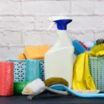 Sponges, plastic brushes and bottles of detergents on a blue wooden table. Household cleaning items on white brick wall surface