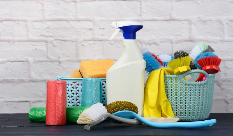 Sponges, plastic brushes and bottles of detergents on a blue wooden table. Household cleaning items on white brick wall surface